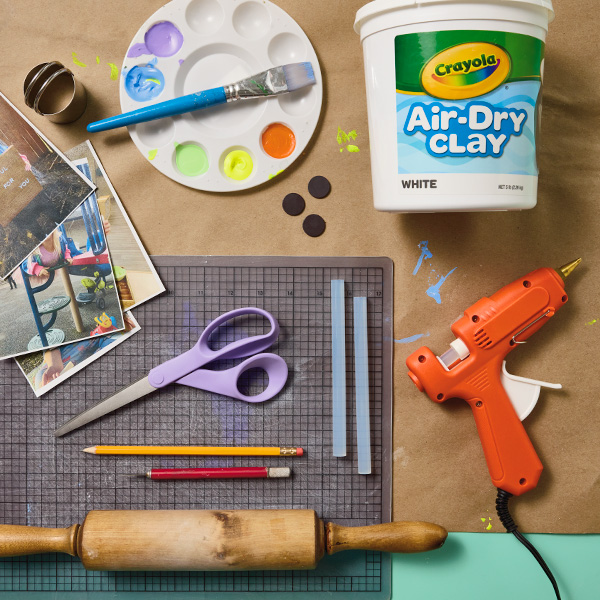 Materials needed to crate the handprint photo frame magnet craft include Crayola air-dry clay, a crafting mat, a hot glue gun, a rolling pin, a pencil, a craft knife, a pair of scissors, a biscuit or circular cookie cutter, magnet disks, a photo of the child making the craft, and paint to decorate with.