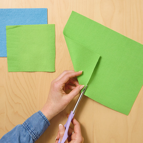 A woman's hands cut a perfect square out of a rectangular sheet of green crafting felt by folding a corner of the felt sheet down to create a right triangle with equal sides and then cutting around it.