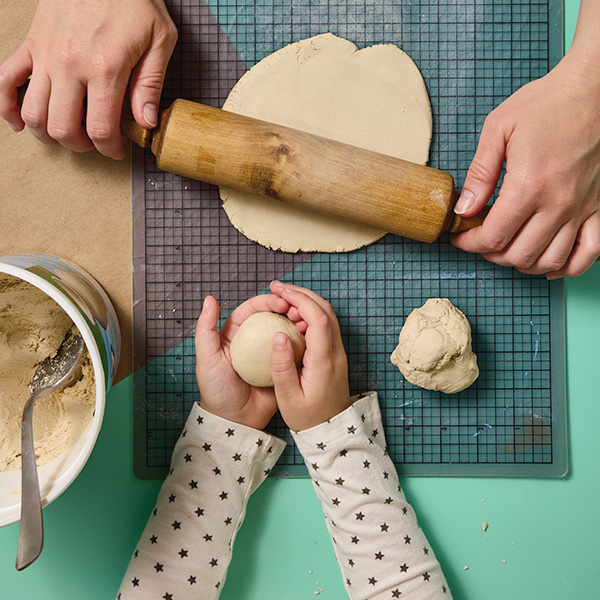 A mother's hands use a small rolling pin to roll a ball of Crayola air-dry clay into a flattened disk on a crafting mat; at the bottom of the frame, a child's hands rest on the opposite end of the crafting mat, holding a ball of the air-dry clay.