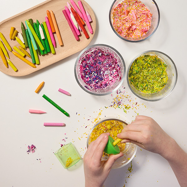 A child uses a pencil sharpener to create shavings from a yellow crayon, which are being collected in a glass bowl sitting underneath the child's hands; a tray holding different shades of yellow, green and pink crayons lays nearby, as well as three additional glass bowls filled with different colors of crayon shavings from the crayons on the tray.