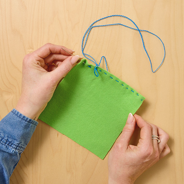 A woman's hands uses an embroidery needle threaded with blue thread to make large, linear stitches around the border of a square of green crafting felt.