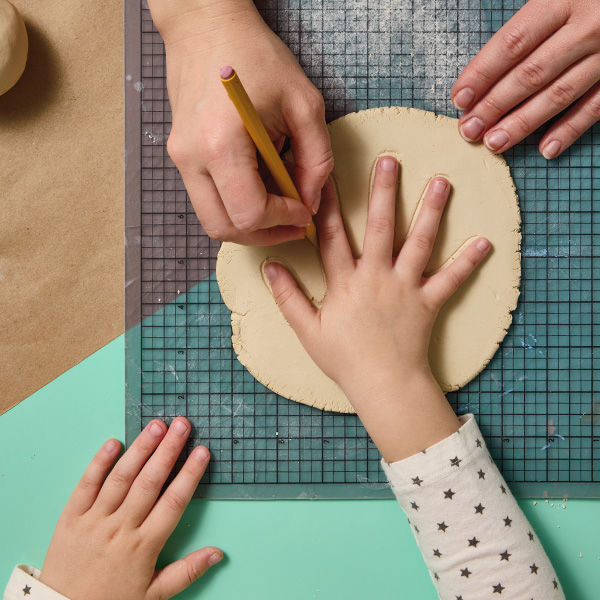 A child places their hand on a flattened piece of air-dry clay with their fingers splayed out; a woman's hand uses a mechanical pencil to trace an outline of the child's hand into the surface of the clay.