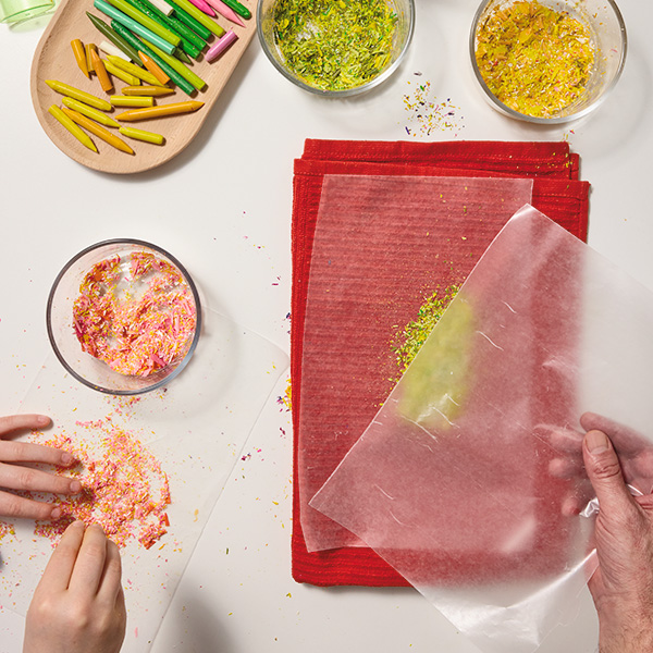 The hands of a child sprinkle crayon shavings onto a piece of wax paper; next to this is a man's hand placing a piece of wax paper on top of his own piece of wax paper with crayon shavings sprinkled on it; the man's wax paper is set on top of a folded kitchen towel, to protect the surface underneath from the heat of a clothing iron used in the next step of the craft.