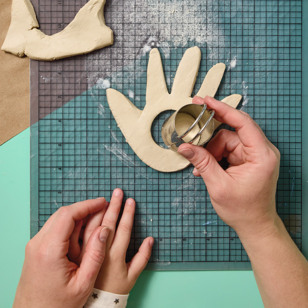 A mom uses her left hand to hold her child's hand, while she uses her right hand to cut a hole into the center of a piece of clay that has been cut from a tracing of the child's hand.