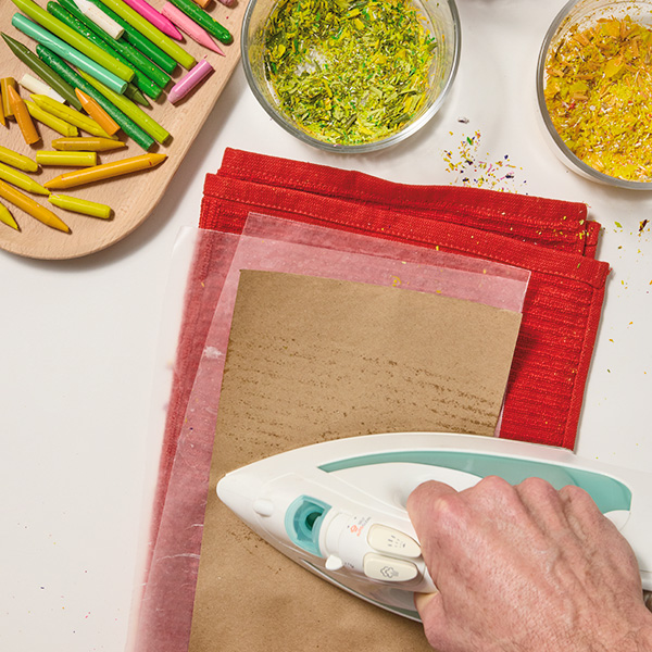 A man uses a clothing iron to melt wax crayon shavings between two pieces of wax paper; a folded kitchen towel has been placed under the wax paper to protect the surface underneath from the heat of the clothing iron; a tray holding crayons in various bright colors and glass bowls holding crayon shavings sit nearby on the same surface.