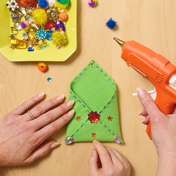 A woman's hands hold a hot glue gun while a child's hand can be seen at the bottom of the frame, reaching in to place a gem embellishment on the felt gift envelope they have made from a piece of crafting felt; nearby is a shallow, yellow dish filled with gems in other shapes and colors, as well as buttons, small trinkets and mini pom-poms.