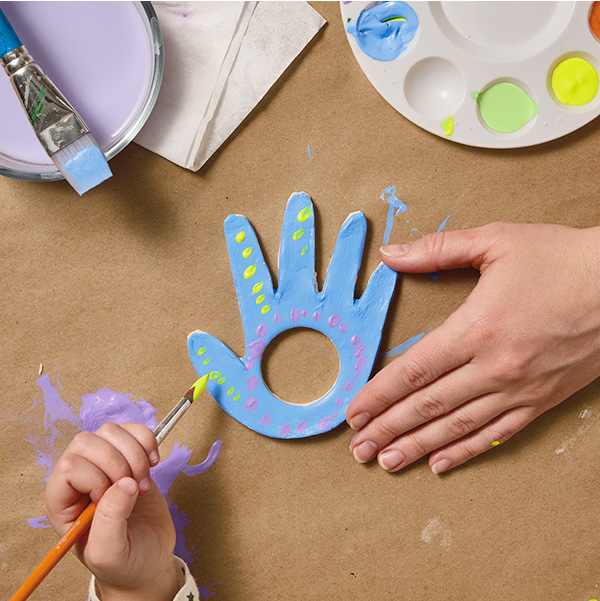 A woman's hand reaches into frame to hold a dried piece of air-dry clay that's been traced and cut out into the shape of a child's hand; there is a perfect circle cut out in the palm of the clay hand; the hand of the child that the craft is modeled after is reaching into frame with a paintbrush to decorate the piece; nearby is a small plastic paint palette with different colors of paint on it.