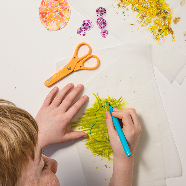 A child uses a marker to draw a shape on the outside of two pieces of wax paper with wax crayon shavings melted in between them; a pair of safety scissors lays nearby to be used in the next step of the craft.