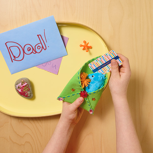 A child's hands place a gift card into a DIY felt gift envelope that they have made with their mother; also in frame is a light yellow tray with a greeting card envelope with 