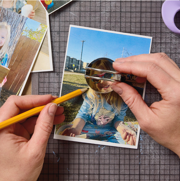 A woman holds a small diameter biscuit cutter down over a photograph of a child with her right hand, while tracing around the biscuit cutter with a pencil using her left hand.