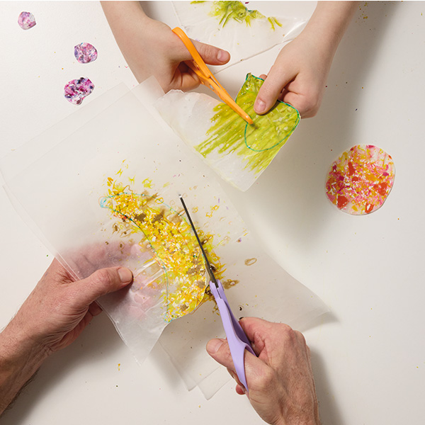A man and a child use scissors to cut shapes out of pieces of wax paper that they have melted colorful wax shavings between; the man is cutting out a shape that looks vaguely like the moon, from yellow wax shavings, while the child cuts out a pear shape from green wax shavings.