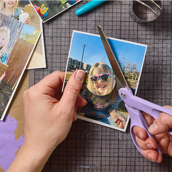 A woman uses a pair of scissors to cut out a circular portion of a printed photograph, featuring her child's face.