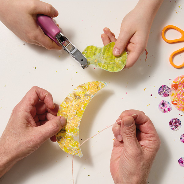 A child uses a hole punch to place a hole in a pear-shaped piece of wax paper with green melted crayon shavings on it; across from him, a man threads a piece of yarn through a hole that has been punched into the yellow, banana-shaped suncatcher he is holding.