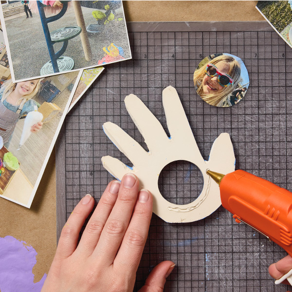 A woman applies hot glue to the back of the air-dry clay that's been traced and cut into the shape of a child's hand with a hole cut in the palm; nearby is a circular photo of a child's face that will be displayed in the frame.