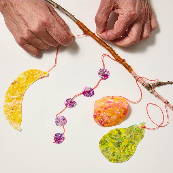 A man ties a melted wax suncatcher made of yellow crayon shavings and in the shape of a banana to a stick with some orange string; other suncatcher shapes have also been tied to the stick using the same method, including a string of purple circles, a big orange and red circle, and a pear-shape suncatcher in different shades of green.