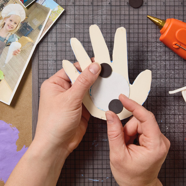 A woman's hands glue magnets to the back of a handprint-shaped photo frame made out of air-dry clay.