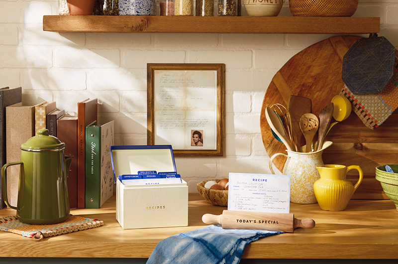 A framed family recipe along with a small photo of the grandmother who wrote it; the framed recipe hangs on a white brick backsplash above a kitchen counter; on the kitchen counter are various cooking tools and recipe books, as well as a recipe box and recipe display stand shaped like a rolling pin.