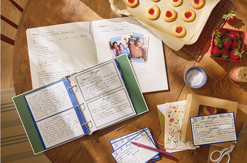 A recipe binder filled with recipe cards sits open on a wooden tabletop; blue and white recipe cards are scattered around the recipe binder, with a family recipe for a thumbprint-style jam cookie written on them; a tray of freshly baked jam-filled cookies sits on the periphery.