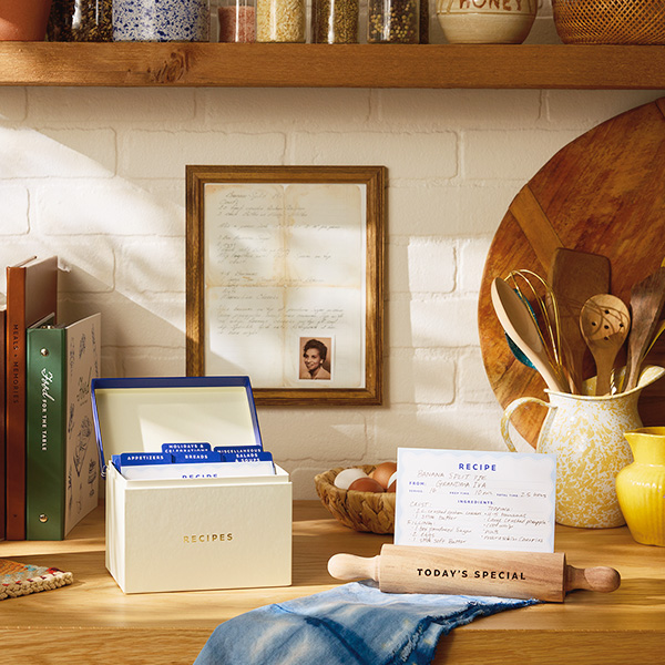 A framed family recipe along with a small photo of the grandmother who wrote it; the framed recipe hangs on a white brick backsplash above a kitchen counter; on the kitchen counter are various cooking tools and recipe books, as well as a recipe box and recipe display stand shaped like a rolling pin.