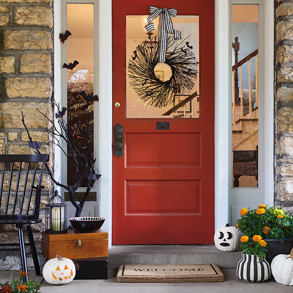 A view of a front porch that has been decorated for Halloween and includes a black front door wreath, black and white jack-o-lanterns positioned on the steps in front of the door, and black bat silhouettes decorating a sidelight to the left of the door.