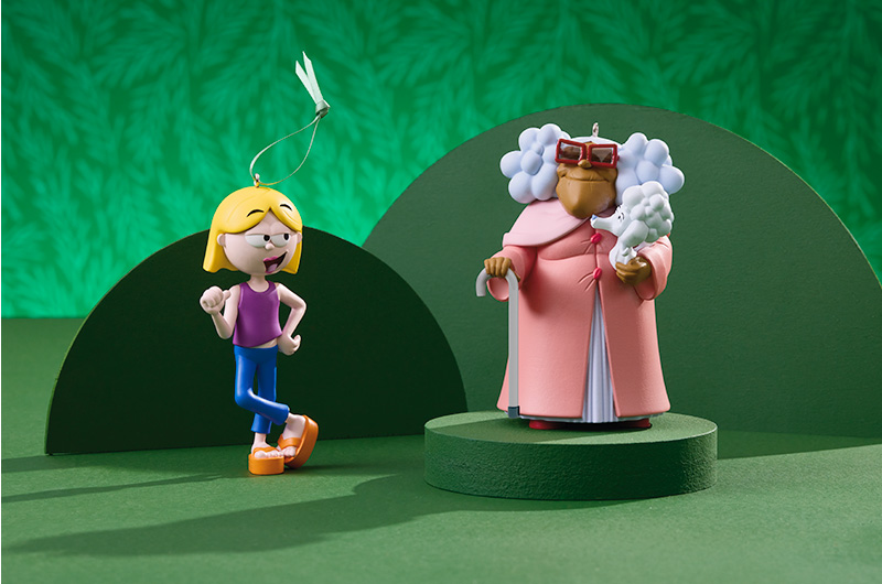 A Lizzie McGuire Keepsake Ornament and a Suga Mama Keepsake Ornament stand near one another on a green surface.