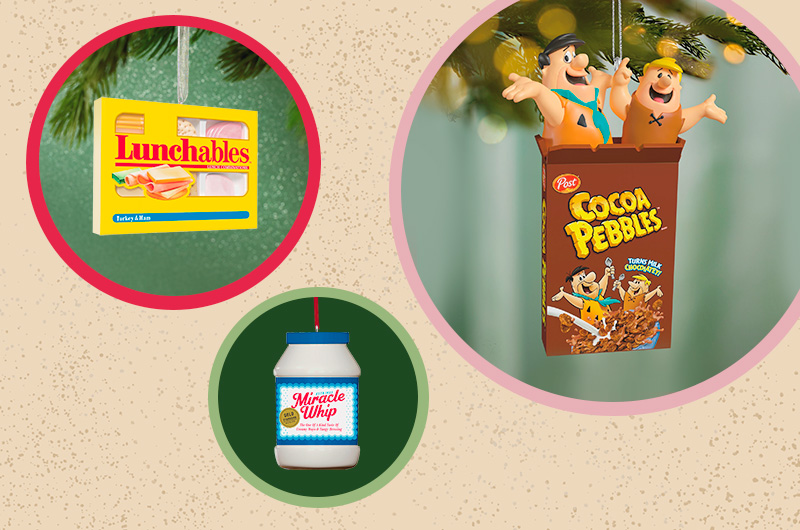 A collection of 90s-nostalgia Keepsake Ornaments collaged on a neutral background, including an ornament that looks just like a Lunchables snack box; an ornament that looks like a jar of Miracle Whip; and an ornament that looks like Fred and Barney from the Flintstones, popping out of a box of Post Cocoa Pebbles.