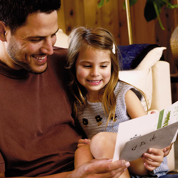 A father sits beside his daughter while holding a Father's Day card and envelope that says 