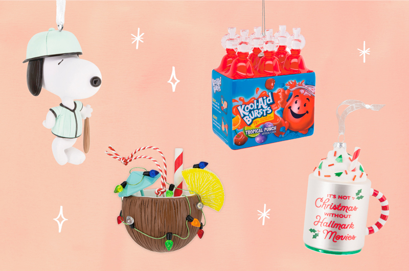 A collection of summer-themed ornaments, including Peanuts Snoopy in a baseball uniform, a tropical drink served in a coconut shell that's been decorated with Christmas lights, an ornament that looks like a six-pack of Kool-Aid squeezie juice bottles, and an ornament that looks like a mug full of hot cocoa with a message on the side that reads, 