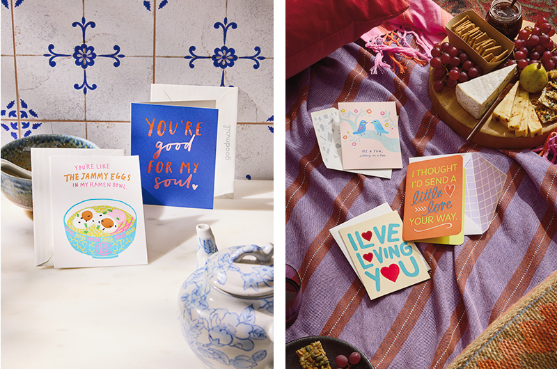 In one lifestyle shot, two cards sit propped up on a kitchen counter top; in another lifestyle shot next to it, a trio of cards are laid out on a blanket next to a charcuterie board, as though part of a floor picnic for an anniversary.