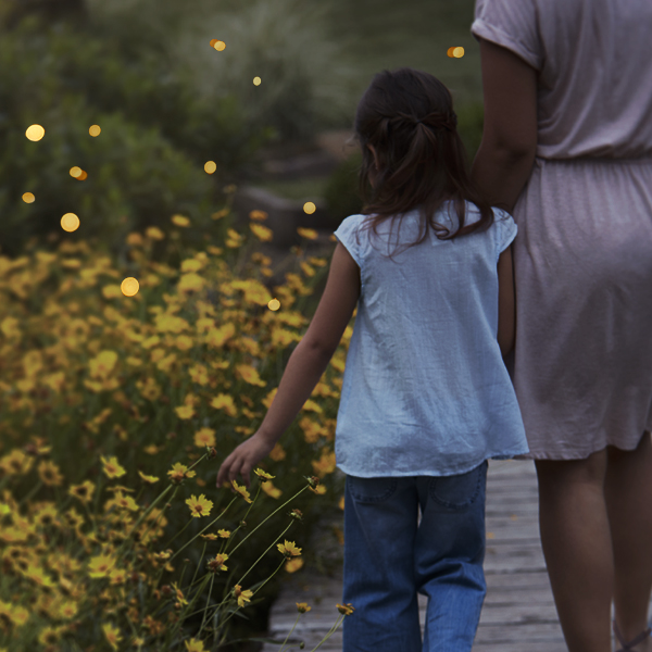 A woman and a little girl walk hand and hand next to a row of yellow wildflowers at dusk; above the wildflowers, fireflies can be seen in flight, their bodies aglow in the evening light.