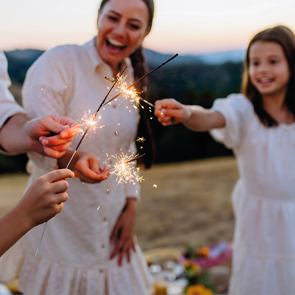 A woman and a young girl, both in white dresses, hold Fourth of July sparklers together to kindle the other's; they are lighting them at dusk on a grassy slope.