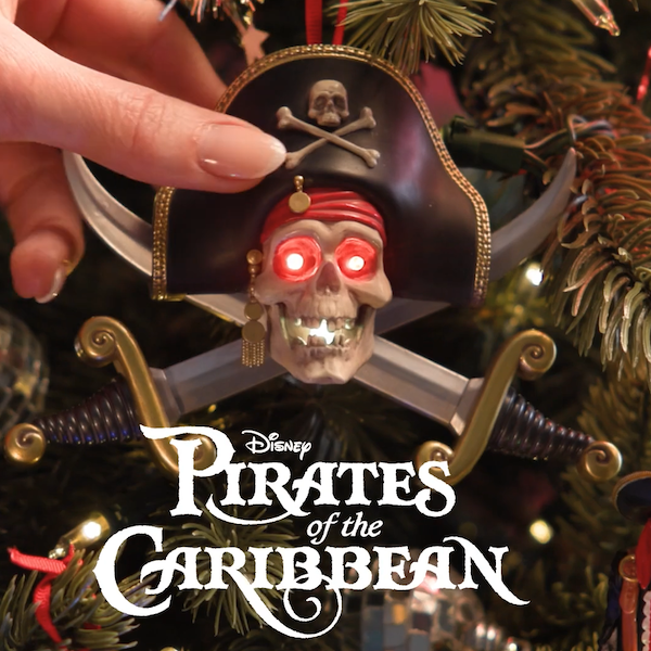 A hand reaches to press a button that activates a Keepsake Ornament hanging on a Christmas tree; the ornament is modeled after the talking skull seen on the Disney Parks Pirates of the Caribbean ride.