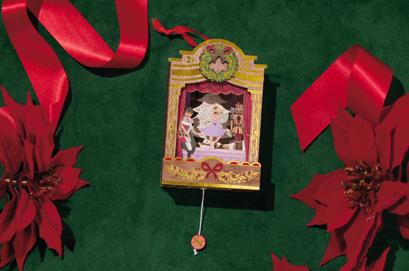 A stunningly detailed Keepsake Ornament depicting a stage production of The Nutcraker ballet; the ornament is made in a layered papercraft style out of laser-cut painted wood, and a pull cord that makes the inner workings of the ornament move.