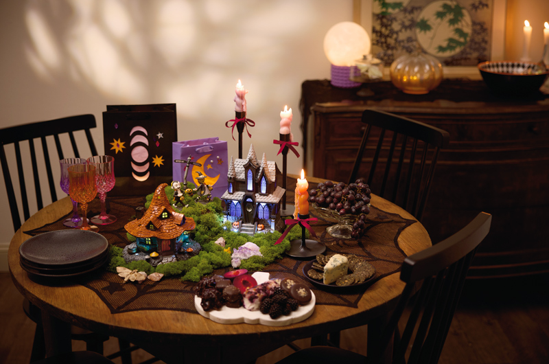 A small, round dining table has been decorated for a witch-themed craft party; at the center of the table are a witch house tabletop decoration and a haunted house tabletop decoration with light, sound and music; surrounding these two figurines are a variety of plated snacks, lit taper candles in black candlesticks, glass drinking goblets in jewel tones, and gift bags that have been transformed into paper lanterns.