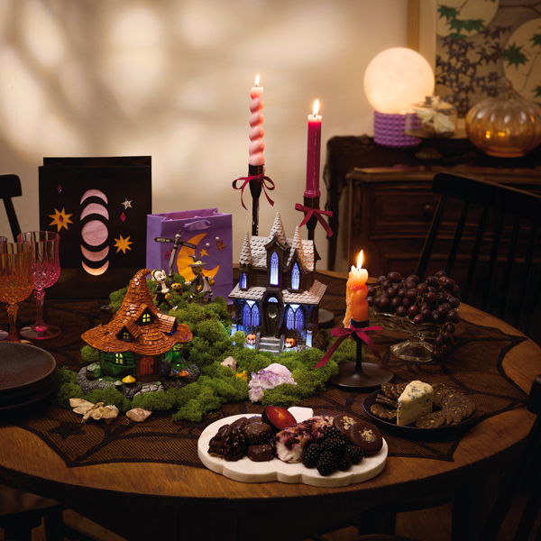 A small, round dining table has been decorated for a witch-themed craft party; at the center of the table are a witch house tabletop decoration and a haunted house tabletop decoration with light, sound and music; surrounding these two figurines are a variety of plated snacks, lit taper candles in black candlesticks, glass drinking goblets in jewel tones, and gift bags that have been transformed into paper lanterns.