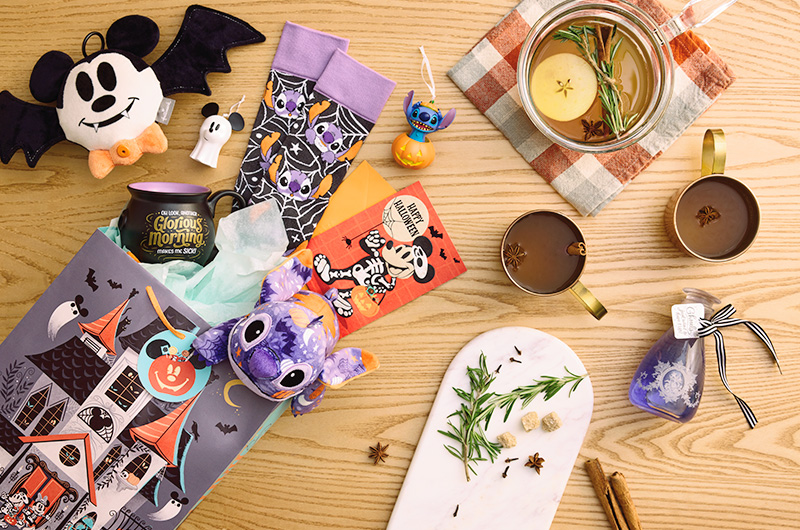 The contents of a Disney-themed Halloween boo bag spilled out onto a tabletop; contents include a plush Stitch covered in a Halloween pattern; a pair of socks covered in a similar pattern featuring Stitch; a plush 