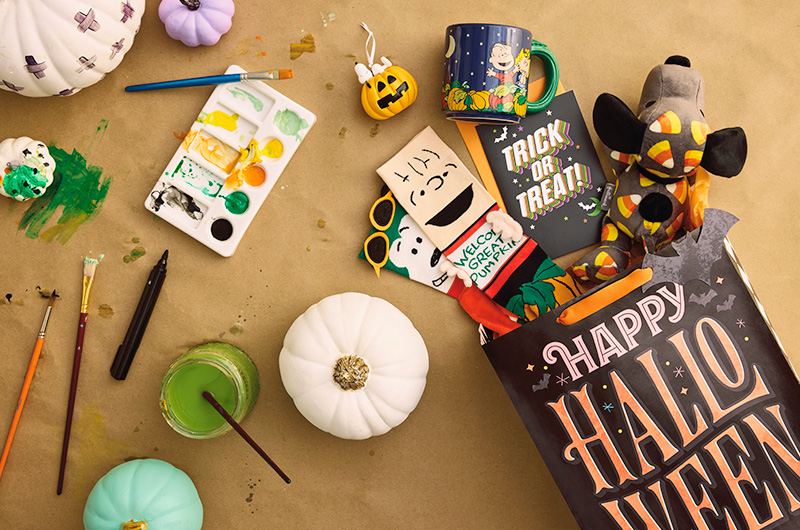 The contents of a Peanuts-themed Halloween boo bag spilled out onto a tabletop; the contents of the boo bag include a plush floppy Snoopy covered in a candy corn pattern; a pair of novelty socks with Linus holding a 