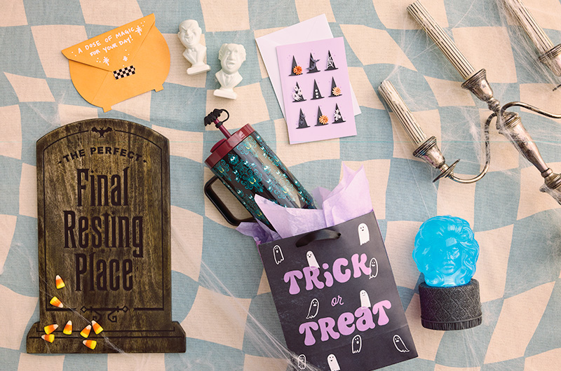 The contents of a Disney The Haunted Mansion Halloween boo bag spilled out onto a table top; contents include a tombstone-shaped charcuterie board; a Madame Leota-shaped water globe with light and sound; an insulated travel mug covered in a pattern reminiscent of the wallpaper in the Haunted Mansion; a pair of Singing Bust salt and pepper shakers; a card with dimensional papercraft witch hats on the front; and an envelope cut into the shape of a cauldron to disguise a gift card with 