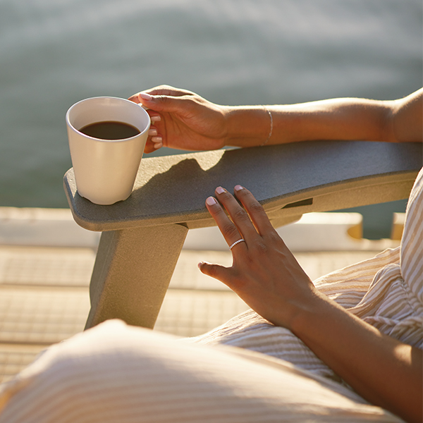 A woman's hands are shown holding a cup of coffee while sitting on a deck chair overlooking a lake.