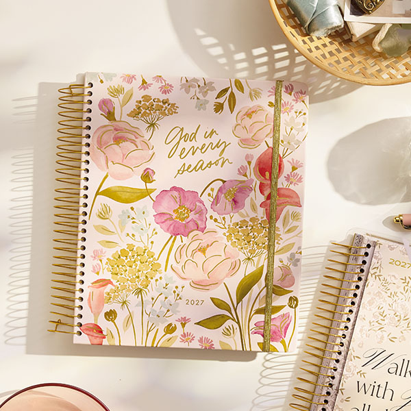 A DaySpring personal planner with a floral, hardback cover and a gold coil binding lays on a white desktop surface.