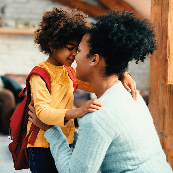 A mother kneels down to hug her son; the son is wearing a backpack, suggesting that he is headed to school.