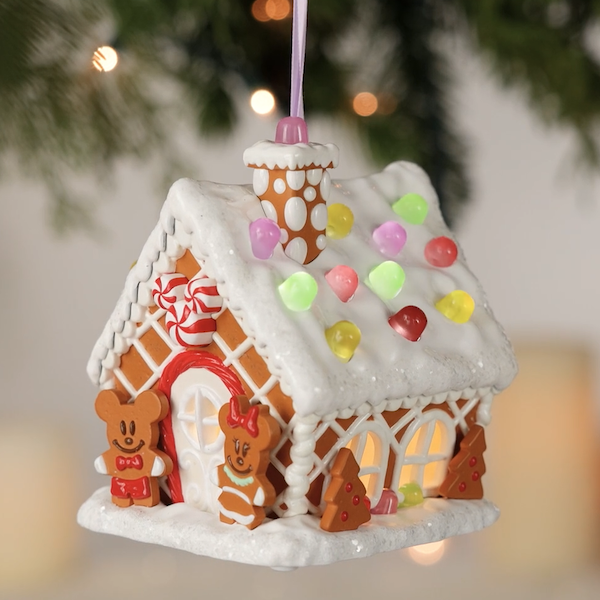 A Keepsake Ornament that looks like a gingerbread house, featuring gingerbread cookie versions of Mickey and Minnie Mouse hangs from a branch on a Christmas tree; the house has realistic looking icing and gumdrops on the roof that light up when a button is pushed; the ornament also plays music.
