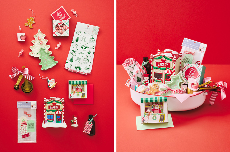 Holiday baking-themed gift basket featuring a ceramic bakery-shaped container, cookie cutters, measuring spoons, baking supplies, kitchen towels, a holiday tea towel, candy canes, and festive Christmas décor arranged against a bright red background.