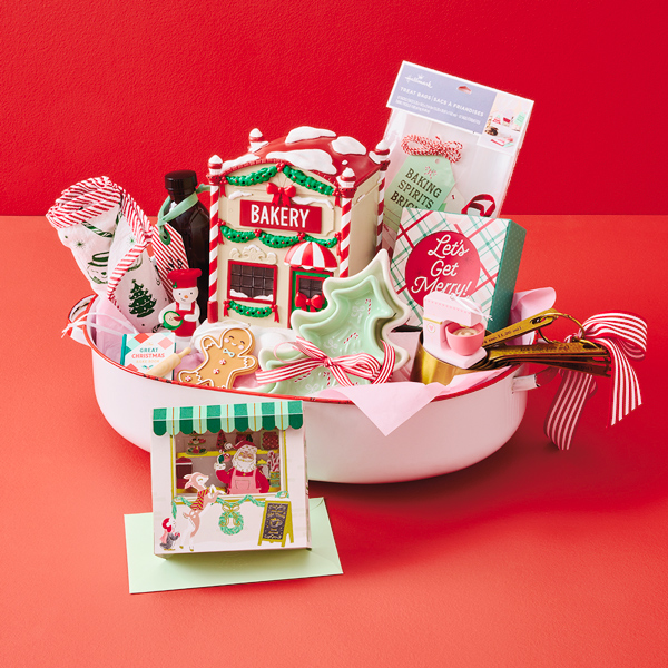 Holiday baking gift basket centered around a ceramic bakery-shaped container, baking accessories, cookie cutters, measuring spoons, kitchen towels, and Christmas treats arranged in a white basin against a red backdrop.