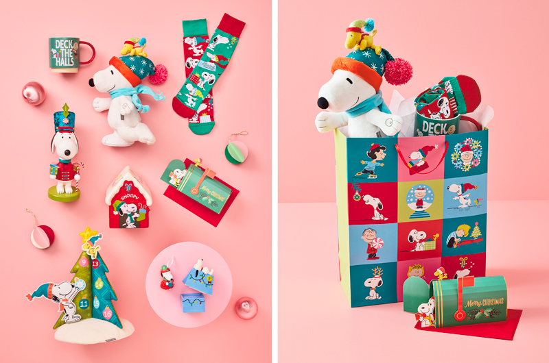 Peanuts-themed Christmas gift basket featuring Snoopy plush toys, holiday socks, ornaments, greeting cards, and festive Peanuts character accessories displayed in a colorful gift bag on a pink background.