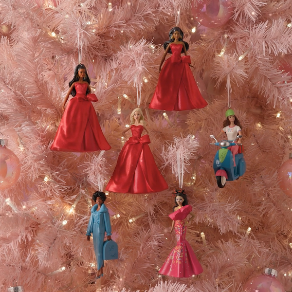 Six Keepsake Ornaments featuring different versions of Barbie hang on a light pink tree; the versions include Caucasian, Black and Latina versions of the 2026 Holiday Barbie, Fashionistas Barbie Doll on Scooter, Barbie Beauty Secrets Christie, and Día De Muertos Barbie.