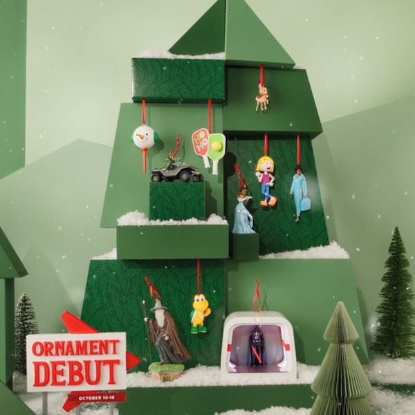 Decorative green Christmas tree display featuring several hanging character and pop-culture ornaments, including a car, wizard figure, turtle character, and miniature figurines, arranged on tiered branches dusted with artificial snow. A red-and-white sign in the lower left reads “Ornament Debut” with smaller text below reading “October 10–18,” while small evergreen trees decorate the background.