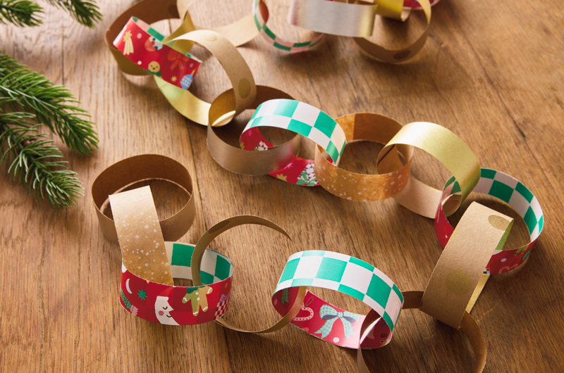 A paper chain made from Christmas gift wrap in different patterns lays draped across a wooden tabletop near an evergreen bough.