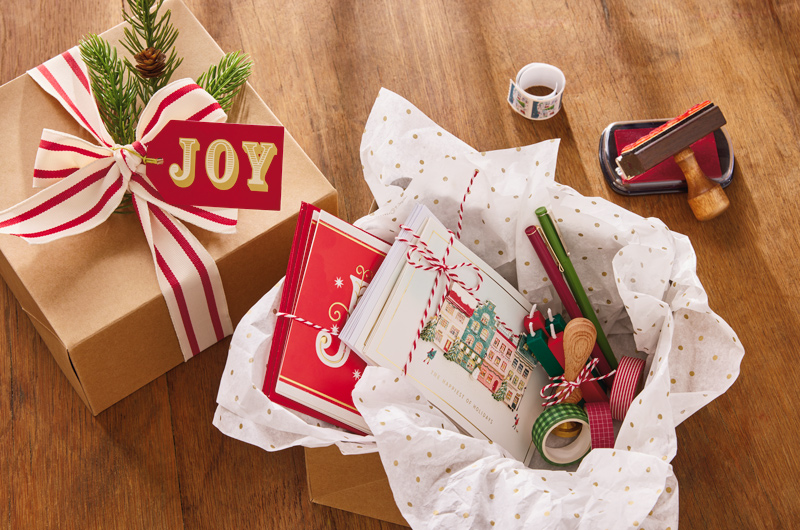 A Christmas card sending kit has been assembled to give as a gift to a loved one; the box includes cards bundled with their envelopes and tied up with red and white striped twine; red and green ink pens; red and green candles for wax seals; a wax seal stamp; and rolls of red and green washii tape in different patterns; the box is lined with white tissue paper speckled with gold polka dots; the lid of the box is decorated with broad, red and white striped ribbon, an evergreen sprig, and a gift tag that reads 