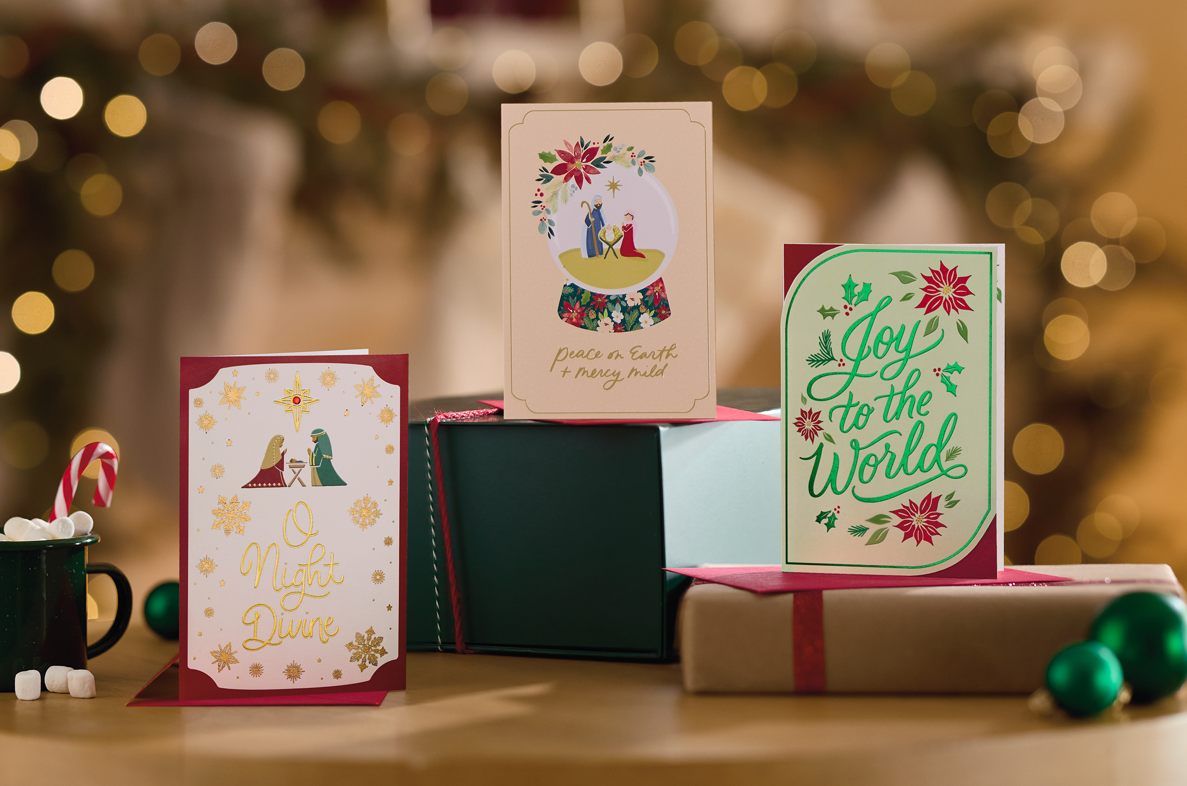 Three Christmas cards featuring scripture and imagery recalling the Nativity stand on a tabletop with gifts and glass ball ornaments.